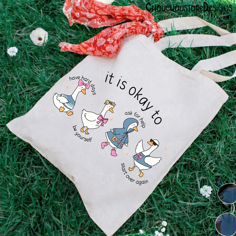 May include: A cream-colored tote bag with the text "it is okay to" and illustrations of geese. The geese are dressed in various outfits, with phrases like "have hard days" and "start over again". The bag has long, cream-colored handles.
