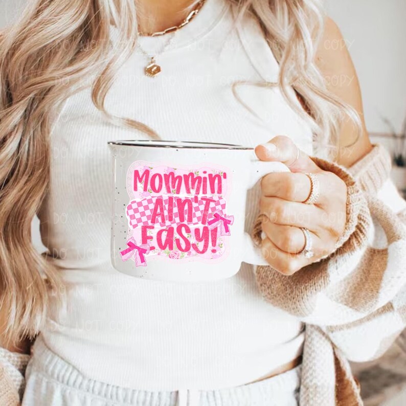 May include: White ceramic mug with a black rim, featuring the text "Mommin' Ain't Easy!" in pink and red lettering. The design includes a checkered pattern and pink bows. The mug is held by a person.