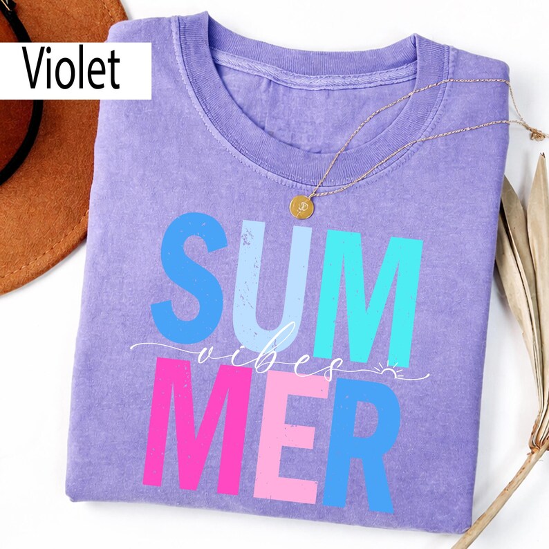 May include: A violet-colored t-shirt with the word "SUMMER" in blue, light blue, and pink, and the words "vibes" and a small sun graphic. A gold necklace with a pendant is on the shirt. A brown hat is visible.