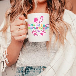 May include: White ceramic mug with the words "EMERGENCY DEPARTMENT" in colorful letters, along with pink bunny ears, paw prints, and hearts. The mug has a handle and is held by a person.