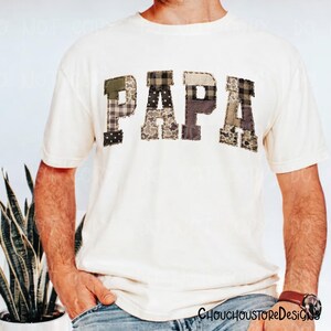May include: Cream-colored t-shirt with the word "PAPA" in patchwork letters. The letters are made of various camouflage and patterned fabrics in shades of green, brown, and gray. A casual, comfortable tee.
