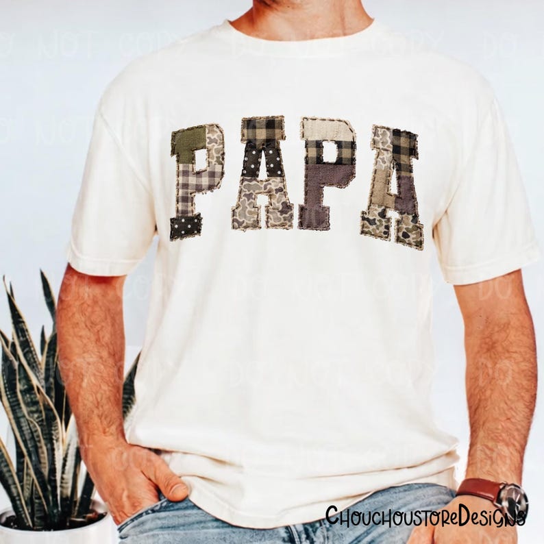May include: Cream-colored t-shirt with the word "PAPA" in patchwork letters. The letters are made of various camouflage and patterned fabrics in shades of green, brown, and gray. A casual, comfortable tee.