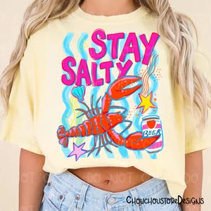 Puede incluir: Camiseta amarilla clara con un dise&ntilde;o gr&aacute;fico colorido. El dise&ntilde;o incluye una langosta, una lata de cerveza y las palabras "STAY SALTY" en rosa. L&iacute;neas onduladas azules, una concha y una estrella completan el dise&ntilde;o.