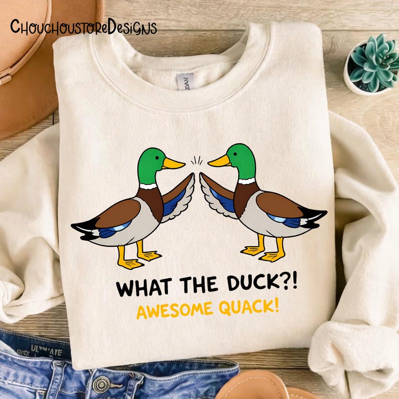 May include: Cream-colored sweatshirt featuring two cartoon ducks with green heads, brown wings, and yellow beaks and feet. The text "WHAT THE DUCK?! AWESOME QUACK!" is printed below the ducks. The brand name "ChouchoustoreDesigns" is printed at the top.