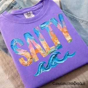 May include: A purple t-shirt with the word "SALTY" in a colorful, textured font. Below the word is a blue wave design. The shirt is folded and resting on a denim surface.