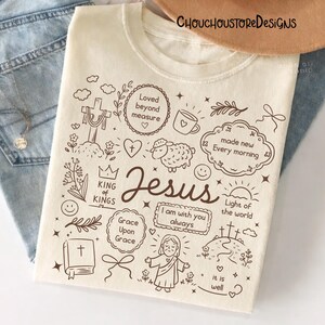 May include: Cream-colored t-shirt with a brown doodle design featuring the word "Jesus" and religious symbols. The design includes a cross, heart, sheep, and the phrases "Loved beyond measure" and "Light of the world."