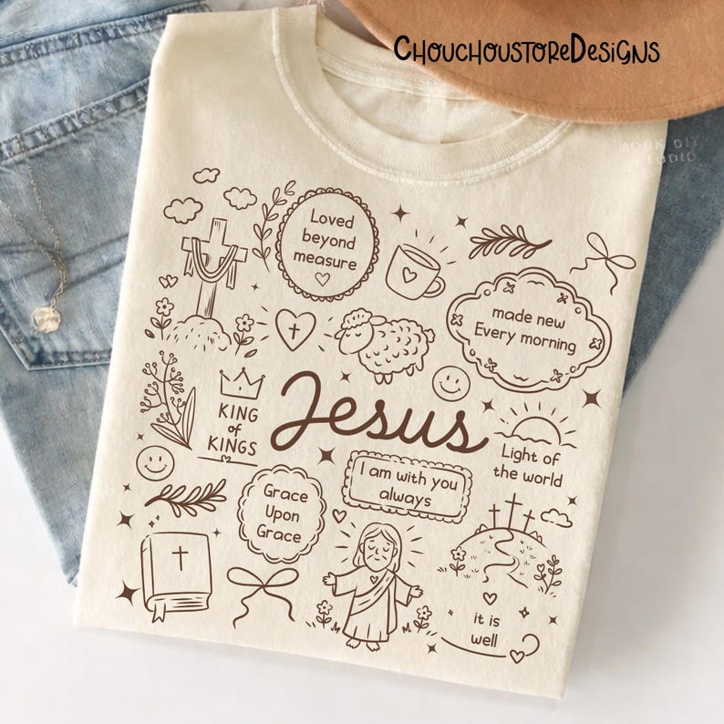 May include: Cream-colored t-shirt with a brown doodle design featuring the word "Jesus" and religious symbols. The design includes a cross, heart, sheep, and the phrases "Loved beyond measure" and "Light of the world."