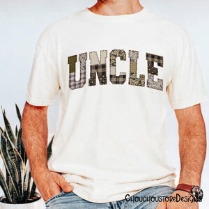 May include: Cream-colored t-shirt with the word "UNCLE" in a patchwork design. The letters feature various patterns, including camouflage and plaid. The shirt has short sleeves and a crew neck.