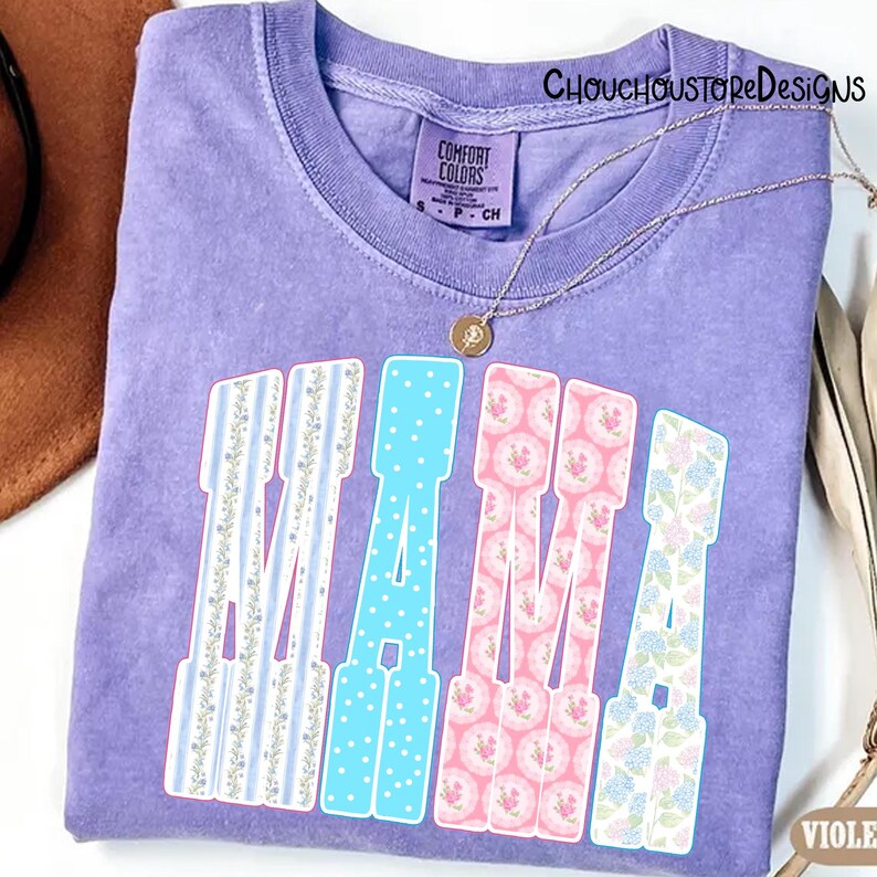 May include: A purple Comfort Colors t-shirt with the word "MAMA" in a decorative font. The letters are patterned with blue stripes, blue polka dots, pink floral, and blue floral designs. A gold necklace is draped across the shirt.