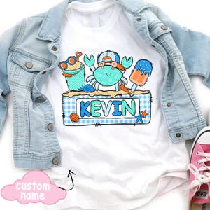 May include: White t-shirt featuring a cartoon crab wearing a hat and sunglasses, with the name "KEVIN" in blue letters. The design includes beach-themed elements like a sand pail, seashell, and popsicle. A light blue denim jacket is draped over the shirt.