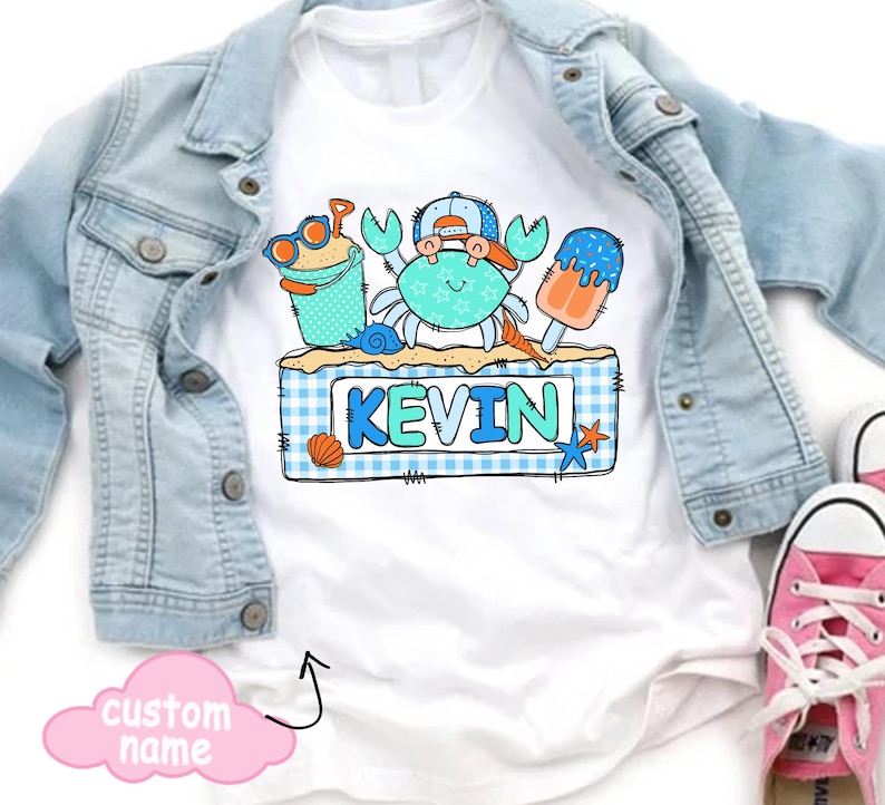 May include: White t-shirt featuring a cartoon crab wearing a hat and sunglasses, with the name "KEVIN" in blue letters. The design includes beach-themed elements like a sand pail, seashell, and popsicle. A light blue denim jacket is draped over the shirt.