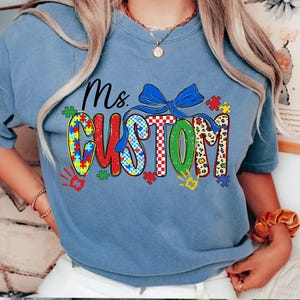 May include: A light blue t-shirt with the words "Ms. CUSTOM" in a colorful design. The word "CUSTOM" features puzzle piece patterns, a blue bow, and handprints. The shirt is a crew neck style.