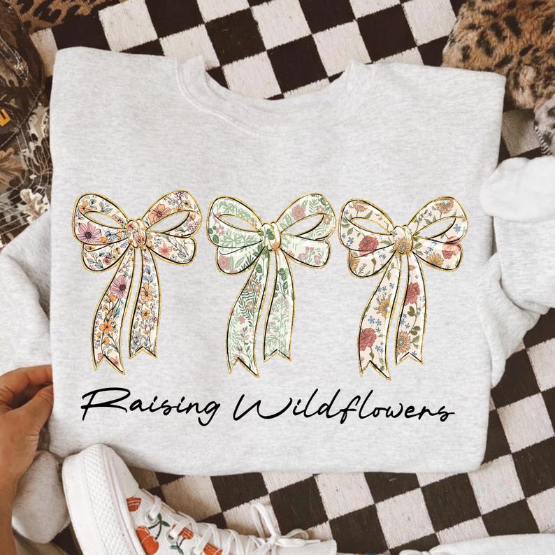 May include: A light gray sweatshirt featuring three floral bow designs with gold outlines. The bows are filled with colorful flowers and greenery. The text "Raising Wildflowers" is printed in black script below the bows.