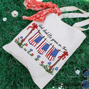 May include: A cream-colored tote bag featuring a patriotic design with three American flags hanging on a clothesline. The flags are red, white, and blue, with the text "God shed His grace on thee." The bag has cream-colored handles and is decorated with flowers.