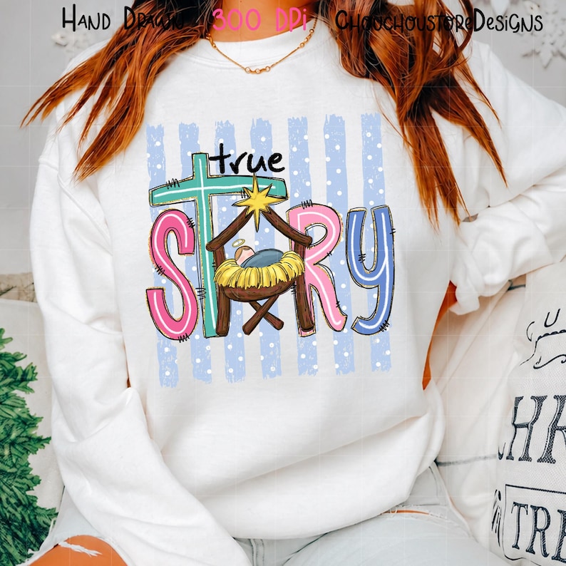 May include: A white long-sleeve sweatshirt featuring a graphic print with the text true story. The design includes a teal cross, a brown manger with a yellow hay bed, and pink and blue letters against a background of light blue vertical stripes.