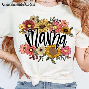May include: Cream-colored t-shirt with the word "Mama" in black script, surrounded by a colorful floral arrangement. The flowers include sunflowers, daisies, and other blooms in shades of yellow, pink, and red. The top is knotted at the waist.