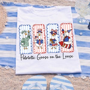 May include: White t-shirt featuring four cartoon geese dressed in patriotic attire. Each goose is framed in red and blue striped borders. The text "Patriotic Goose on the Loose" is printed below the geese. The shirt is on a blue and white striped towel.
