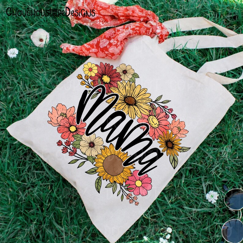 May include: A white canvas tote bag with the word "Mama" in black script, surrounded by a colorful floral design featuring sunflowers and other flowers in shades of yellow, pink, and orange. The bag has long, cream-colored handles.