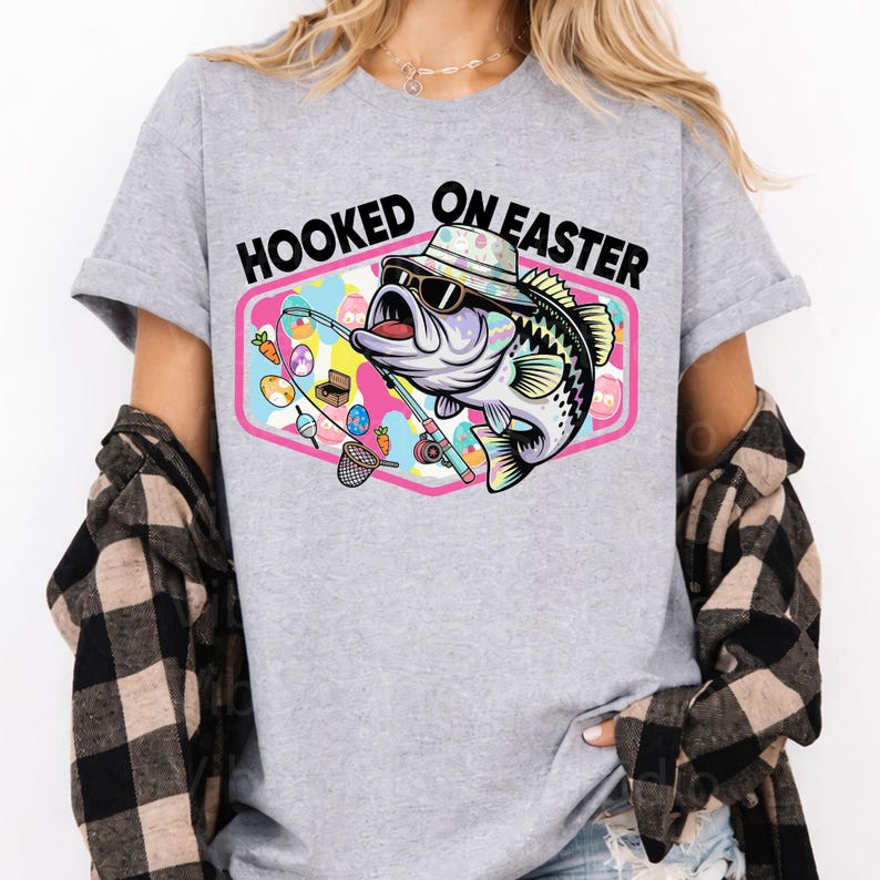 Pode incluir: Camiseta cinza claro com um gr&aacute;fico de um peixe usando &oacute;culos de sol e chap&eacute;u, segurando uma vara de pescar. O gr&aacute;fico est&aacute; dentro de um hex&aacute;gono rosa e branco com as palavras "HOOKED ON EASTER".