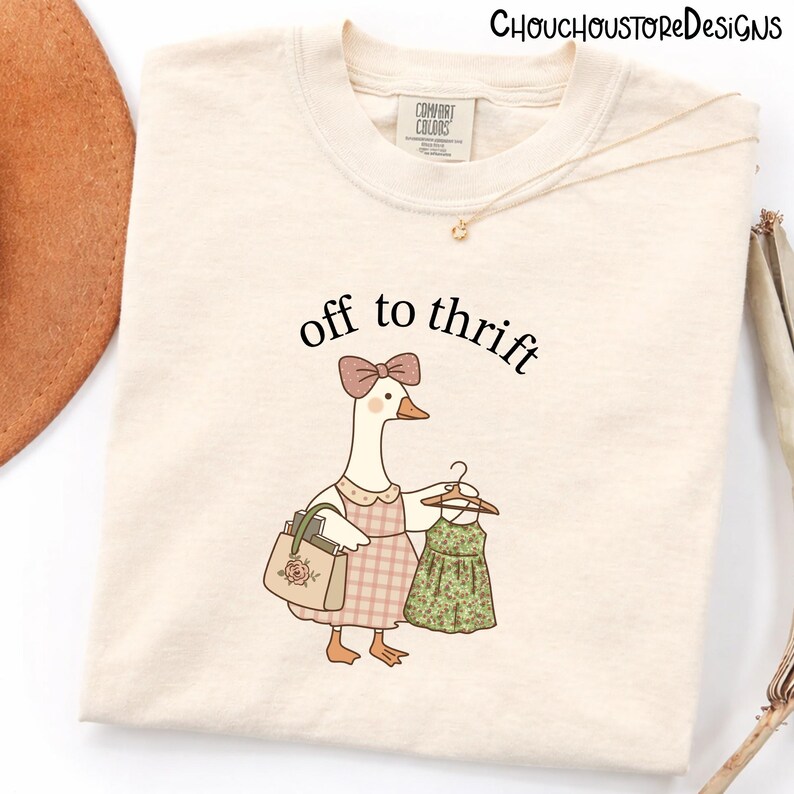 May include: Cream-coloured t-shirt featuring the text "off to thrift" above a cartoon goose wearing a dress and holding a dress on a hanger. The goose carries a purse with a rose design. The shirt has a Comfort Colors tag.