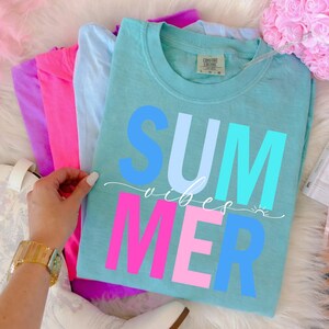 May include: A stack of colorful t-shirts, including teal, pink, purple, and light blue. The teal shirt features the words "SUMMER vibes" in blue and pink block letters. A hand with a gold watch is visible.