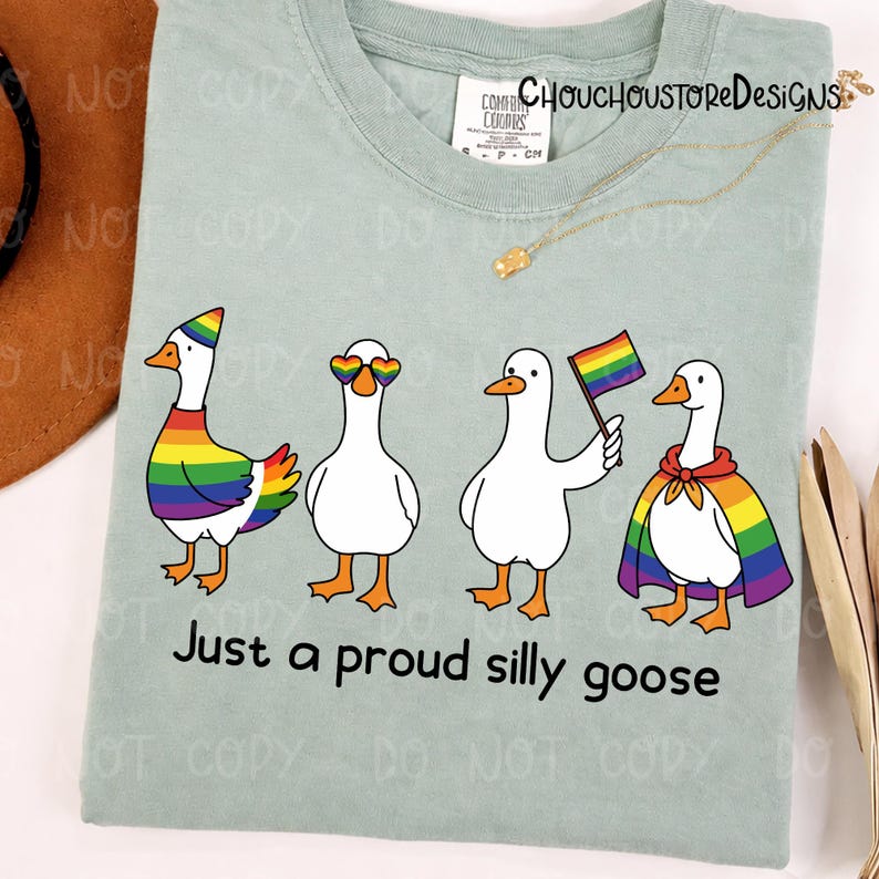 K&ouml;nnte beinhalten: Hellgr&uuml;nes T-Shirt mit vier Cartoon-G&auml;nsen mit Regenbogen-Accessoires. Der Text "Just a proud silly goose" ist darunter aufgedruckt. Das T-Shirt hat ein Comfort Colors-Etikett.
