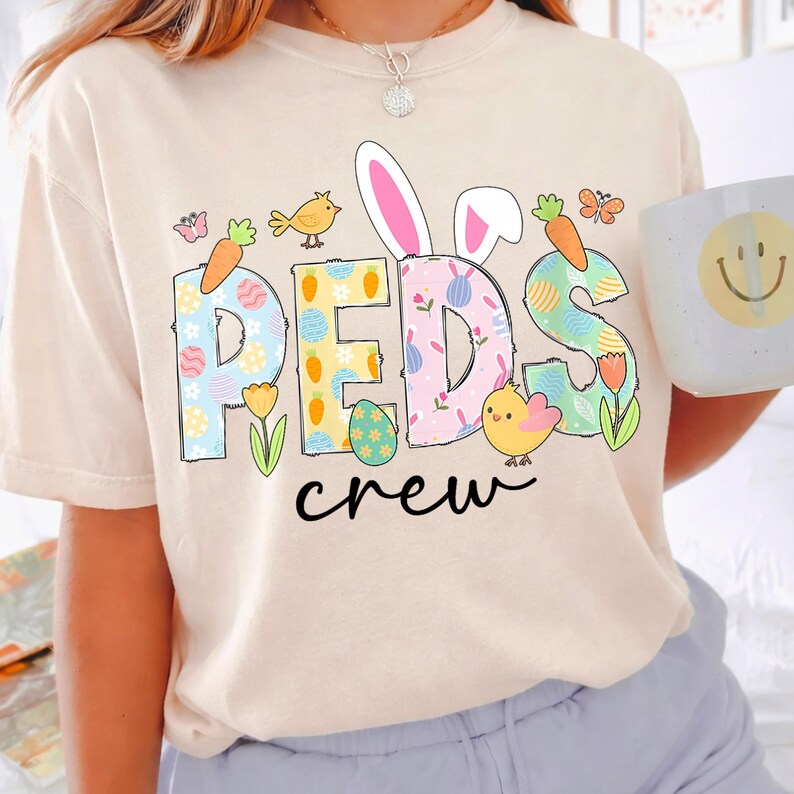 May include: Beige t-shirt with the words "PEDS crew" in colorful, Easter-themed lettering. The letters are decorated with carrots, eggs, flowers, and bunny ears. A yellow chick and a bird are also featured.