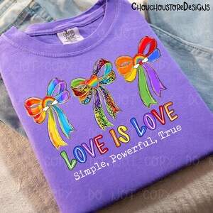 K&ouml;nnte beinhalten: Ein lila T-Shirt mit drei bunten Schleifen mit Regenbogen-Akzenten und dem Text "LOVE IS LOVE". Die Schleifen haben Regenbogen- und Leopardenmuster. Darunter steht "Simple, Powerful, True".