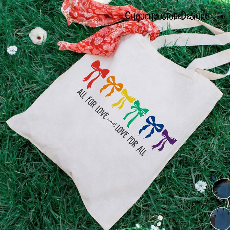 May include: A white tote bag with a rainbow of colorful bows and the text "ALL FOR LOVE and LOVE FOR ALL". The bag has long, off-white handles and is lying on green grass. A red scarf is draped over the bag.