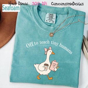 May include: A seafoam green t-shirt featuring a graphic of a white goose wearing a pink bow, holding a coffee cup and a pink notebook labeled Lesson Plans with a pencil. The text Off to teach tiny humans is printed in white above the goose.