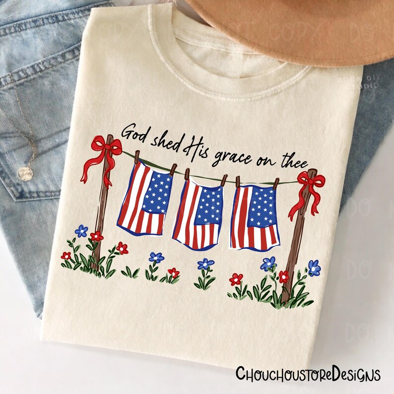 May include: Cream-colored t-shirt featuring a patriotic design. The graphic includes American flags hanging on a clothesline, with the text "God shed His grace on thee." Red bows and flowers add decorative elements. The brand name "ChouchoustoreDesigns" is at the bottom.
