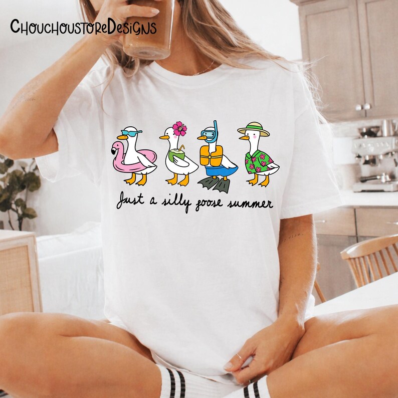 May include: White t-shirt featuring a whimsical illustration of four cartoon geese, each accessorized for summer fun. One wears sunglasses and a flamingo floatie, another a flower, a third has snorkel gear, and the last a Hawaiian shirt. The text "Just a silly goose summer" is below.