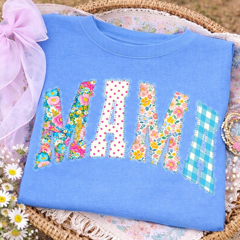 May include: A pale blue sweatshirt featuring the word "MAMA" in appliqu&eacute;d letters. The letters are crafted from diverse patterned fabrics, including floral designs, polka dots, and a blue and white gingham check. The sweatshirt is presented in a woven basket.
