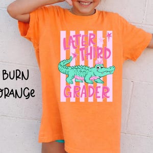 May include: Burn orange t-shirt with the text "Later Third Grader" in pink, featuring a cartoon alligator. The alligator is green with pink accents and a pink bow. The shirt is a short-sleeved crew neck.