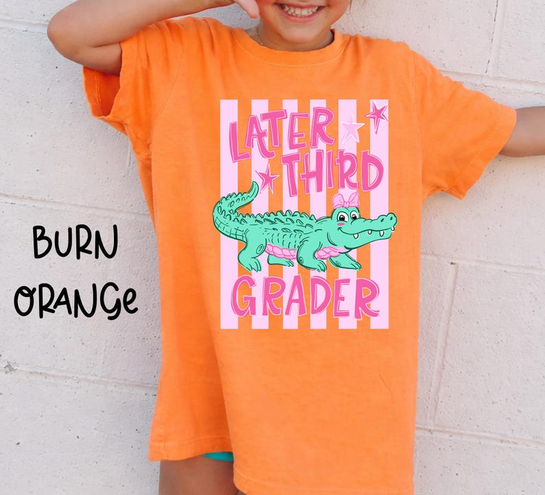May include: Burn orange t-shirt with the text "Later Third Grader" in pink, featuring a cartoon alligator. The alligator is green with pink accents and a pink bow. The shirt is a short-sleeved crew neck.