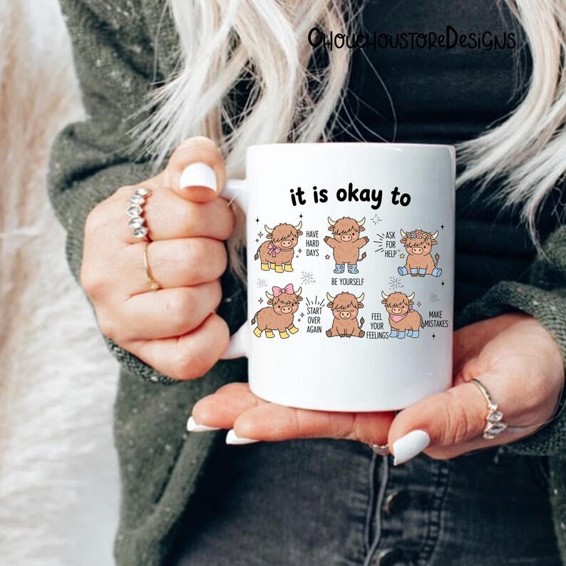 May include: White ceramic mug featuring the phrase "it is okay to" in black, with illustrations of cartoon Highland cows. The cows are depicted with various accessories and text bubbles with positive affirmations.