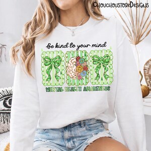 May include: White sweatshirt with a graphic design promoting mental health awareness. The design includes green bows, a brain with flowers, and the text "Be kind to your mind" and "Mental Health Awareness".