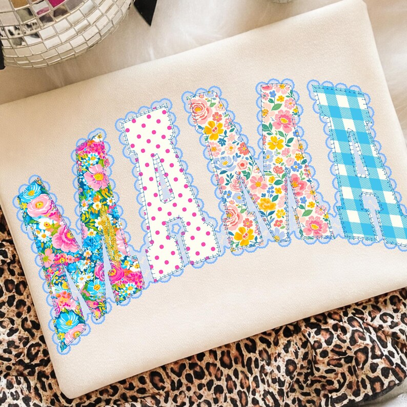 May include: Cream sweatshirt featuring the word "MAMA" appliqu&eacute;d in diverse floral and patterned fabrics. The letters are outlined with a light blue scalloped trim. The fabric patterns include pink polka dots and blue gingham.