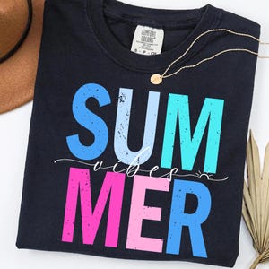 May include: Navy blue t-shirt with the words "SUMMER VIBES" in large, colorful, distressed lettering. The word "SUM" is blue, "MER" is pink, and the word "VIBES" is in white script. A gold necklace is draped across the shirt.
