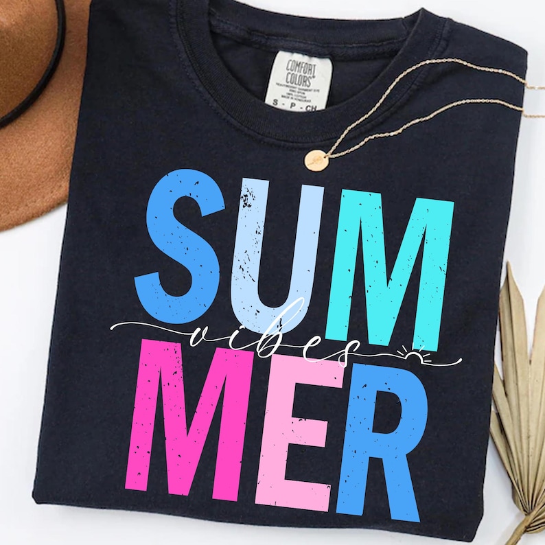 May include: Navy blue t-shirt with the words "SUMMER VIBES" in large, colorful, distressed lettering. The word "SUM" is blue, "MER" is pink, and the word "VIBES" is in white script. A gold necklace is draped across the shirt.
