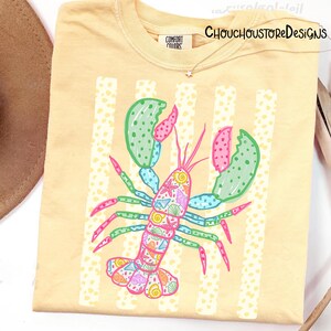 May include: A light yellow Comfort Colors t-shirt featuring a colorful lobster illustration. The lobster is decorated with seashells and other marine life. Vertical white stripes with yellow polka dots are behind the lobster. The text "CHOUCHOUSTOREDESIGNS" is at the top right.