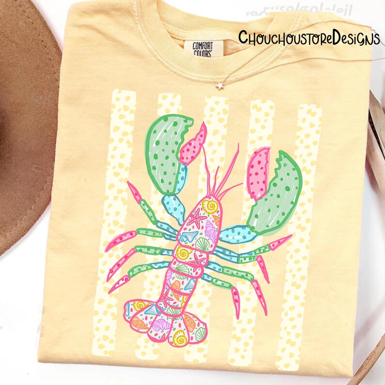 May include: A light yellow Comfort Colors t-shirt featuring a colorful lobster illustration. The lobster is decorated with seashells and other marine life. Vertical white stripes with yellow polka dots are behind the lobster. The text "CHOUCHOUSTOREDESIGNS" is at the top right.