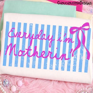 Pu&ograve; includere: Borsa tote bianca con strisce verticali blu e la scritta rosa "Everyday I'm Motherin'". Un fiocco rosa &egrave; sul lato destro. Lo sfondo presenta tessuti piegati in rosa, verde e giallo.