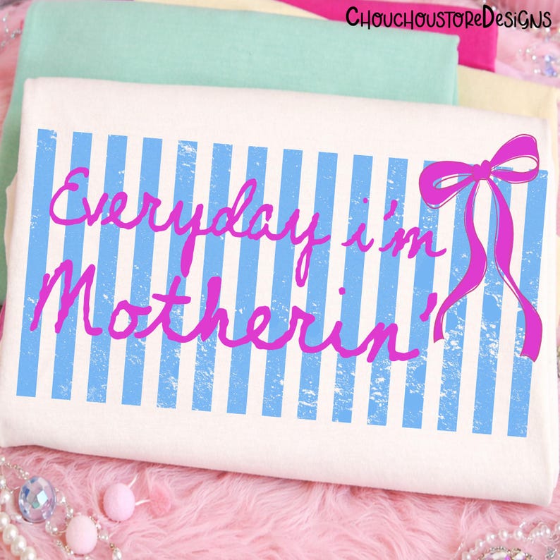 Pu&ograve; includere: Borsa tote bianca con strisce verticali blu e la scritta rosa "Everyday I'm Motherin'". Un fiocco rosa &egrave; sul lato destro. Lo sfondo presenta tessuti piegati in rosa, verde e giallo.