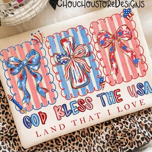 May include: A cream-colored graphic tee featuring patriotic designs. The shirt displays three framed illustrations of bows and a cross, all in red, white, and blue. The text "GOD BLESS THE USA LAND THAT I LOVE" is printed below the illustrations.