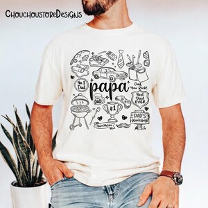 May include: Cream-colored t-shirt with a black graphic design featuring the word "papa" and various illustrations like a car, grill, and tools. The shirt also includes text that says "No. 1 Dad", "Best Dad Ever", and "Dad's Workshop".