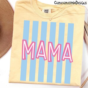 Puede incluir: Una camiseta amarilla clara con rayas verticales azules y la palabra "MAMA" en letras rosas con contornos blancos. La camiseta tiene cuello redondo y una peque&ntilde;a etiqueta que dice "COMFORT COLORS".