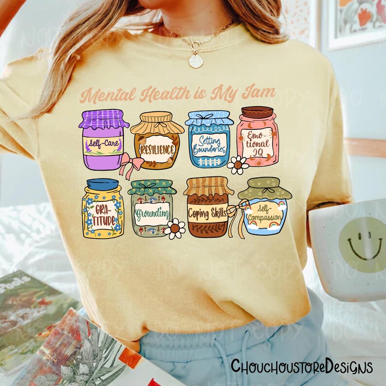May include: A light yellow t-shirt with the text "Mental Health is My Jam" and illustrations of jam jars labeled with self-care concepts. The jars are in various colors and designs, including "Self-Care", "Resilience", and "Gratitude".