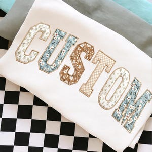 Peut inclure: Coussin blanc avec le mot "CUSTOM" brodé dans un style universitaire. Les lettres présentent des motifs floraux et à carreaux dans des tons de bleu, marron et beige, avec une bordure dorée. Le coussin est posé sur une surface à carreaux noirs et blancs.
