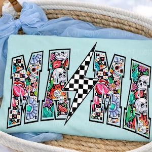 May include: A light blue t-shirt with the word "MAMA" in a bold, outlined font. The letters are filled with colorful illustrations of skulls, flowers, dice, and lips. A black and white checkered lightning bolt is in the center.